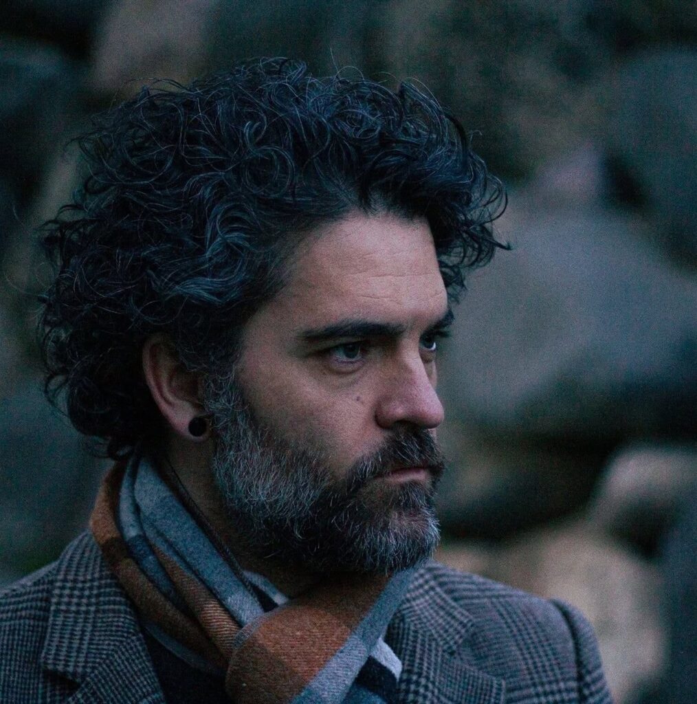 A closeup image of Vincent Blackwood, a middle aged man with curled hair and a salt and pepper beard, wearing a scarf and dress coat.